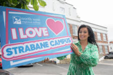 Woman in a colourful dress outside a large building holding a big sign that says I love Strabane campus