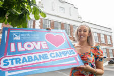 Woman in a colourful dress outside a large building holding a big sign that says I love Strabane campus