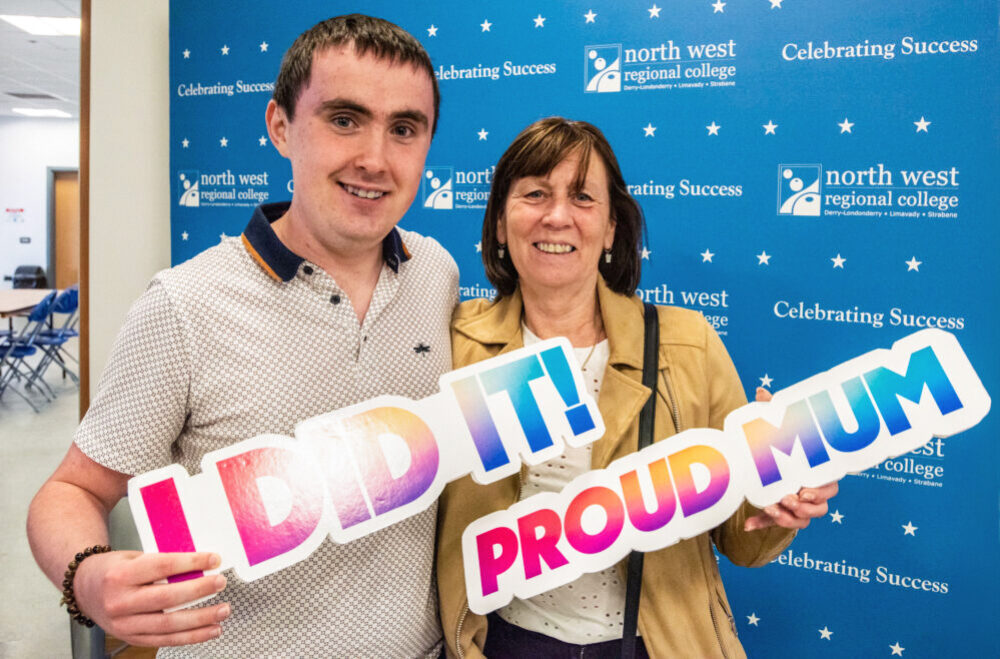 A male and female are in front of blue signage with NWRC written on it, The male is holding an I did it sign while the female is holding a Proud mum sign
