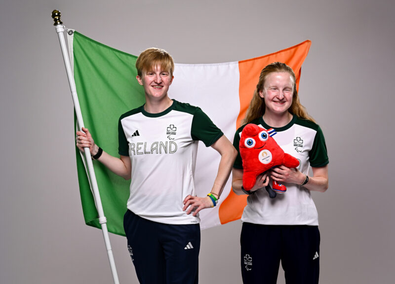 Two young women in sports tops holding irish flag and red Paralympics mascot plushie