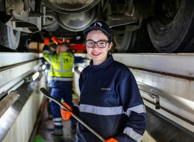 Female motor vehicle apprentice smiles at camera with mechanic worry on lorry in background