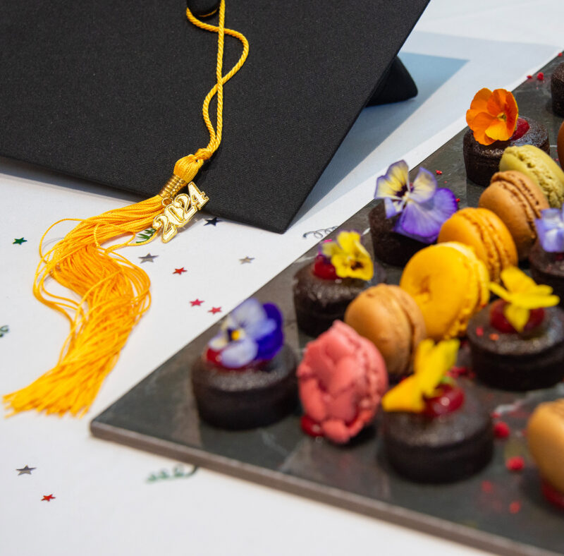 Graduation mortar board with yellow tassel sitting on a white table with colourful desserts