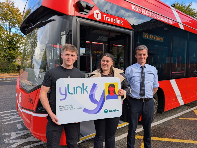 Two students and a bus driver outside standing outside with a bus behind them. They are hold a card with ylink on it