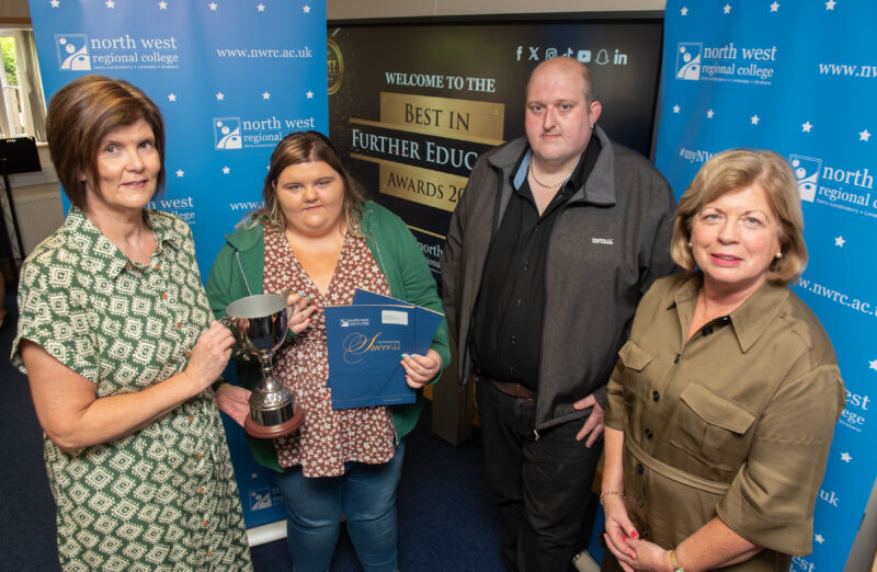 Female student is presented with a trophy by another female. There is also a man and another lady in the photo. They are all standing in front of blue branding for NWRC