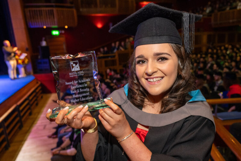 Woman holding a trophy wearing a graduation gown and cape. She is standing in front of a crowd in an auditorium.