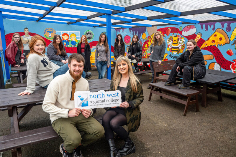 Large group of females and one male in a cafe which has been painted with a mural. They are holding a logo that says NWRC.