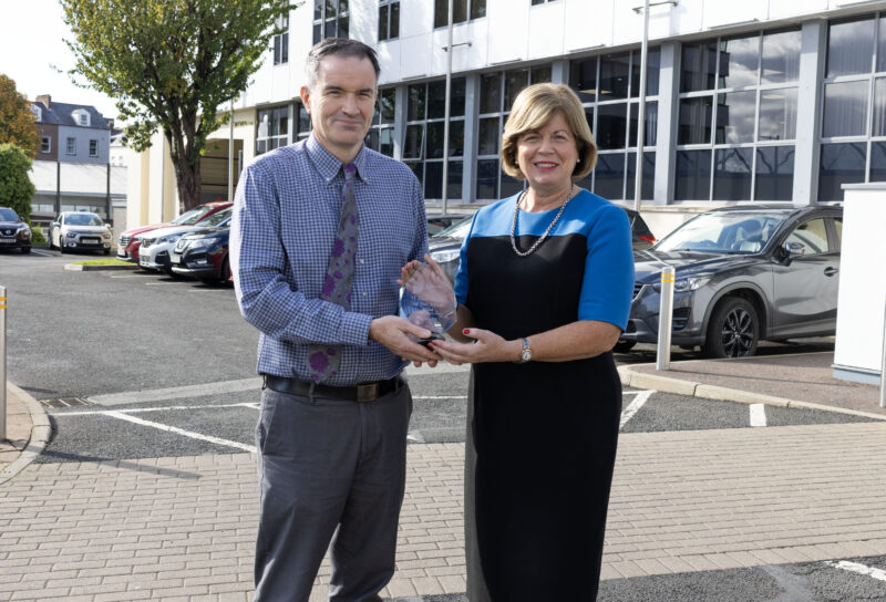 Man and woman outside a college building. They are smiling and holding a crystal award.