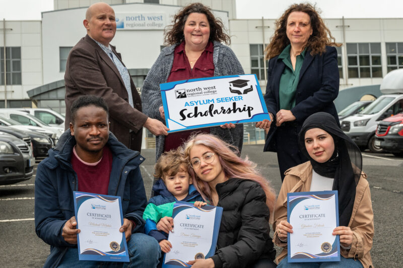 seven people standing in two rows outside a college building with cars parked at both sides. The back row of three people are holding a sign 'NWRC Asylum Seeker Scholarship' and the front row are holding certificates.