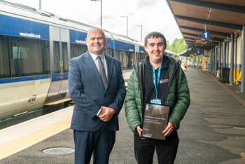 A man in a suit, and a student in a hoodie on the platform of a railway station with a train behind them.