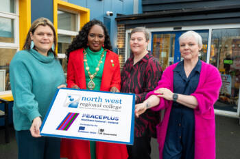 Four women outside a community centre hold a graphic with the NWR and Peaceplus logo