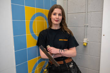 Student smiles, she is holding tiling tool and leaning against coloured tiles