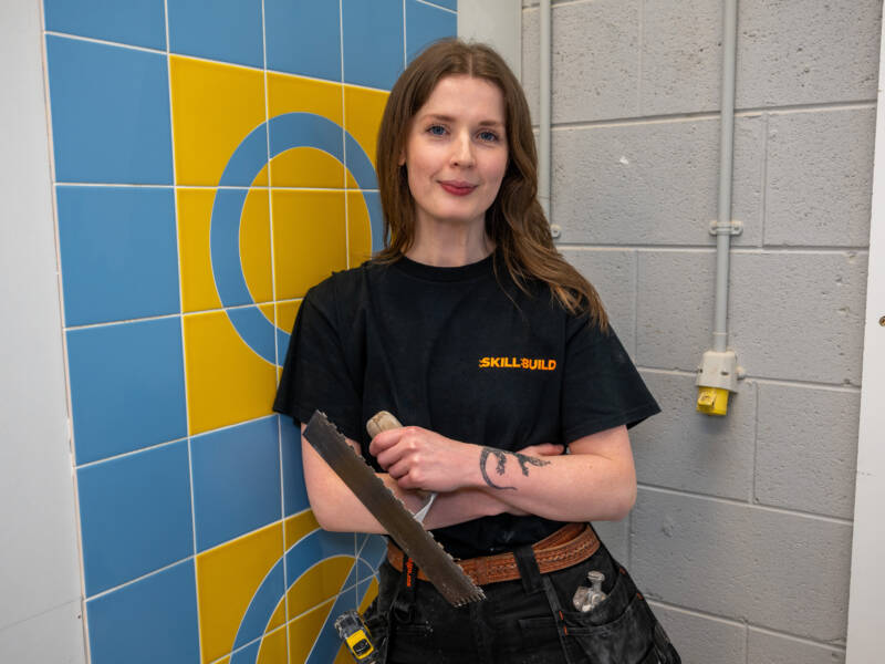 Student smiles, she is holding tiling tool and leaning against coloured tiles