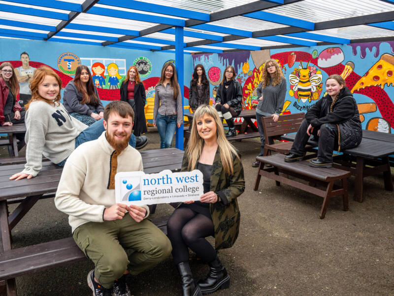 Large group of females and one male in a cafe which has been painted with a mural. They are holding a logo that says NWRC.